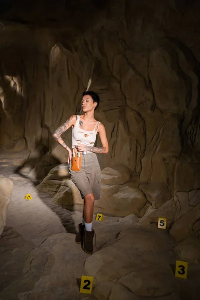 Comprimento total de arqueólogo sexy no topo do tanque em pé perto de cartões numerados na caverna — Stock Photo