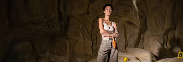 Joven arqueólogo morena en pantalones cortos y camiseta de pie con los brazos cruzados en la cueva oscura, pancarta - foto de stock