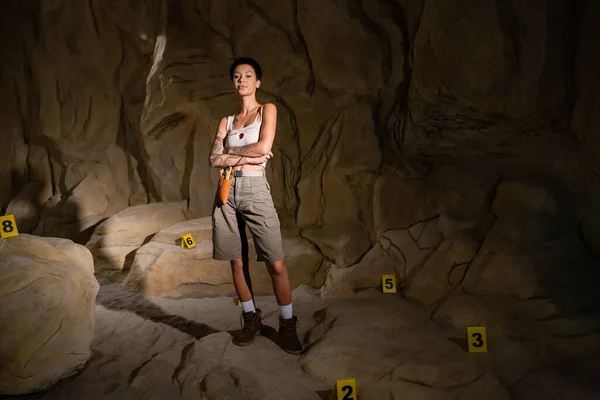 Comprimento total de jovem arqueólogo de pé com braços cruzados perto de marcas numeradas na caverna — Fotografia de Stock