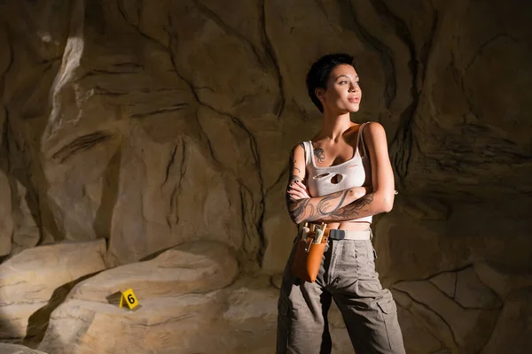 Young and sexy archaeologist in tank top standing in cave with crossed arms and looking away — Stock Photo