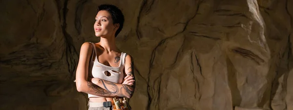 Tattooed archaeologist in sexy tank top standing with crossed arms and looking away in cave, banner — Stock Photo