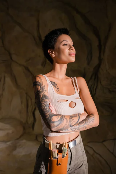 Sexy and tattooed archaeologist standing with crossed arms and looking away in dark cave — Stock Photo