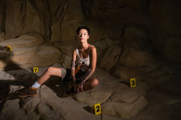 Junger Archäologe in Tank-Top und kurzer Hose, der in die Kamera blickt, während er in der Nähe nummerierter Markierungen in der Höhle sitzt — Stockfoto