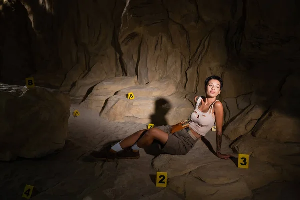 Joven arqueólogo morena sentado cerca de tarjetas numeradas en la cueva oscura y mirando hacia otro lado - foto de stock