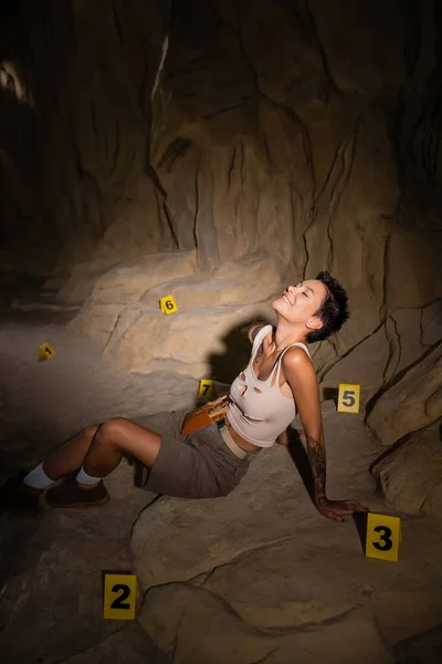 Arqueólogo feliz y sexy sentado cerca de tarjetas numeradas en la cueva oscura - foto de stock