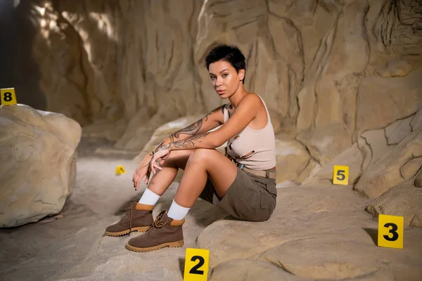 Young tattooed archaeologist sitting near numbered cards in cave and looking at camera — Stock Photo
