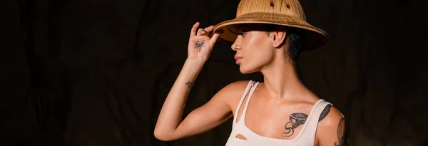 Jeune archéologue tatoué en débardeur ajustant le chapeau de safari dans le désert sombre, bannière — Photo de stock