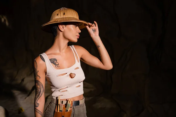 Tattooed archaeologist with tools in waist bag adjusting safari hat while looking away in dark desert — Stock Photo