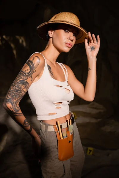 Sexy tattooed archaeologist in tank top touching safari hat and looking at camera in dark cave — Stock Photo