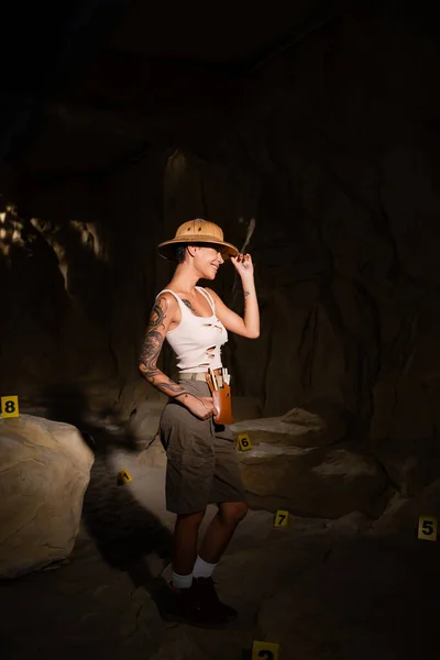 Seitenansicht eines jungen und glücklichen Archäologen, der neben nummerierten Karten in der Höhle steht und Safari-Hut berührt — Stockfoto