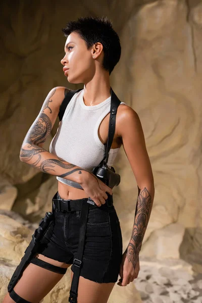 Sexy archaeologist in white crop top and black shorts taking gun out of holster in cave — Stock Photo