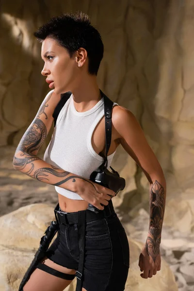 Tattooed archaeologist with short hair taking gun out of holster in cave — Stock Photo