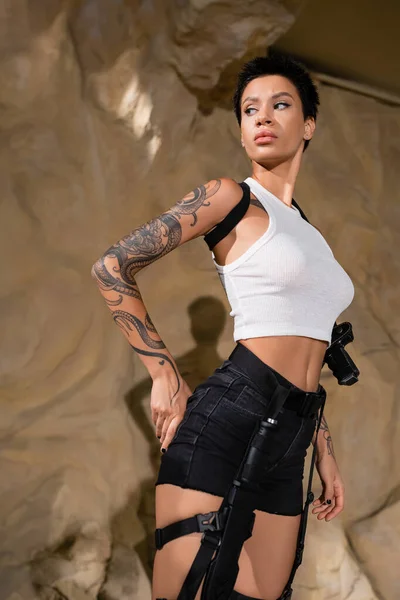 Serious tattooed archaeologist in white crop top and black shorts standing with hand on hip in desert — Stock Photo