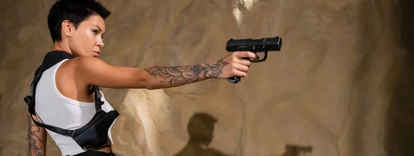Вид збоку татуйованого археолога з кобуром, який тримає пістолет у пустелі, банер — стокове фото