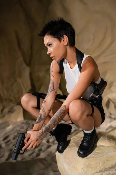 Sexy morena arqueóloga en blanco crop top sosteniendo pistola en piedra en cueva - foto de stock