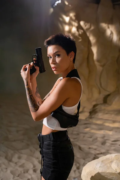 Morena arqueóloga con funda de pistola y mirando a la cámara en el desierto - foto de stock