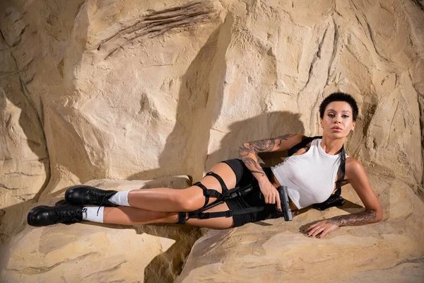 Full length of sexy archaeologist in shorts and crop top looking at camera while lying with gun near rock — Stock Photo