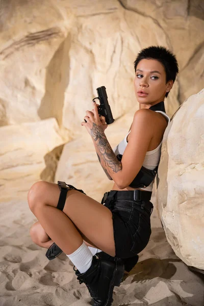 Sexy archaeologist with short hair holding gun while hiding behind rock in cave — Stock Photo