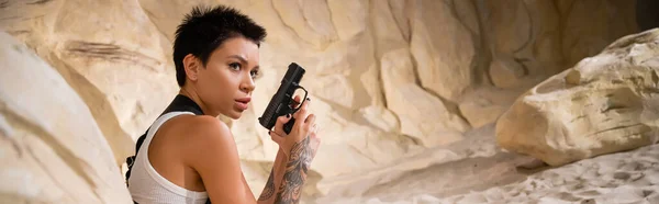 Arqueólogo tatuado con pistola de pelo corto mientras se esconde detrás de la roca en la cueva, pancarta - foto de stock