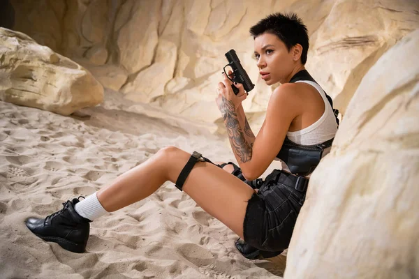 Sexy archaeologist with short hair and tattoo holding gun while hiding behind rock in cave — стоковое фото