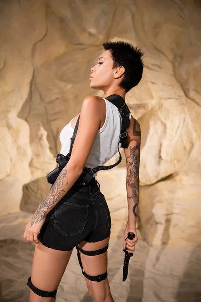 Tatuado e sexy arqueólogo em shorts com coldre segurando faca afiada — Fotografia de Stock