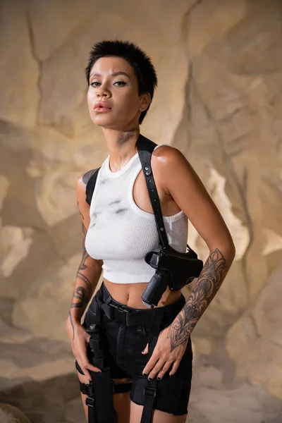 Tattooed young archaeologist with dirty crop top standing with gun in holster — Stock Photo
