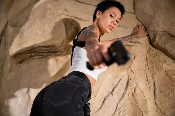 Низкий угол обзора татуированного молодого археолога в сексуальном наряде, нацеленного с пистолетом на размытый передний план — стоковое фото