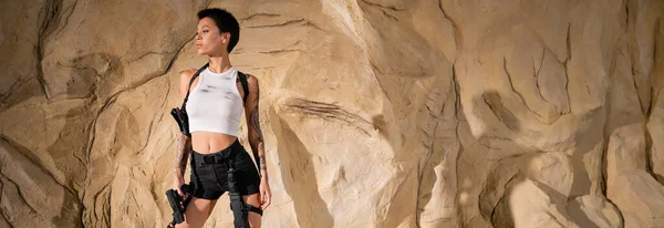 Joven arqueólogo tatuado en traje sexy sosteniendo pistola y de pie cerca de las rocas, pancarta - foto de stock