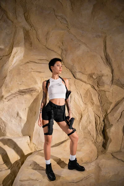 Full length of tattooed archaeologist in sexy outfit holding gun and standing near rocks — Stock Photo