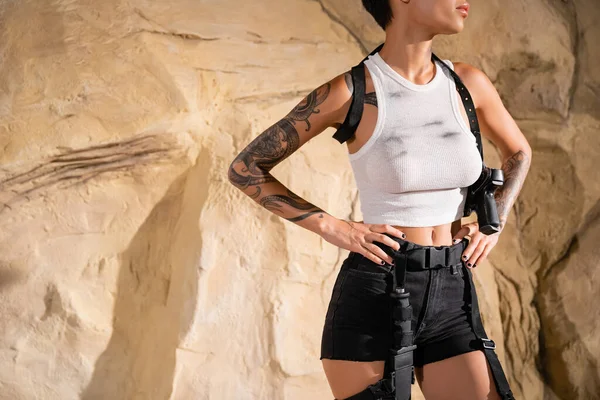 Ausgeschnittene Ansicht eines bewaffneten und tätowierten Archäologen im sexy Outfit, der mit den Händen auf Hüften in der Nähe von Felsen steht — Stockfoto