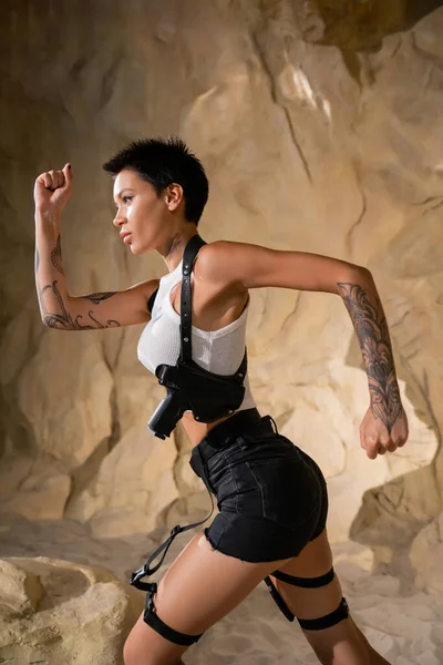 Side view of armed and tattooed archaeologist in sexy outfit running in cave — Stock Photo