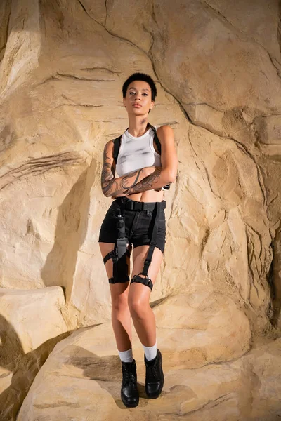 Piena lunghezza di archeologo tatuato e armato in abito sexy in piedi con le braccia incrociate vicino rocce — Foto stock