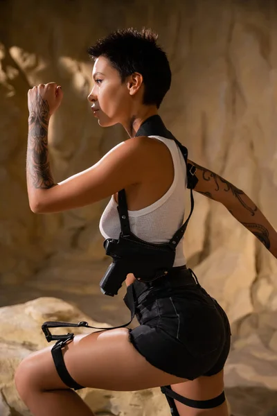 Profile of armed and tattooed archaeologist in sexy outfit running in cave — Stock Photo
