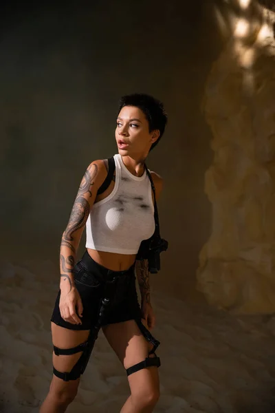 Curioso y tatuado arqueólogo en traje sexy con pistola en funda caminando en cueva - foto de stock