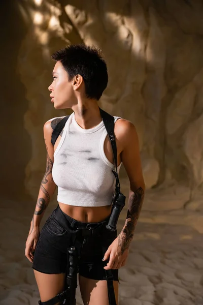 Tatuado arqueólogo no sexy roupa com arma no coldre olhando para longe na caverna — Fotografia de Stock
