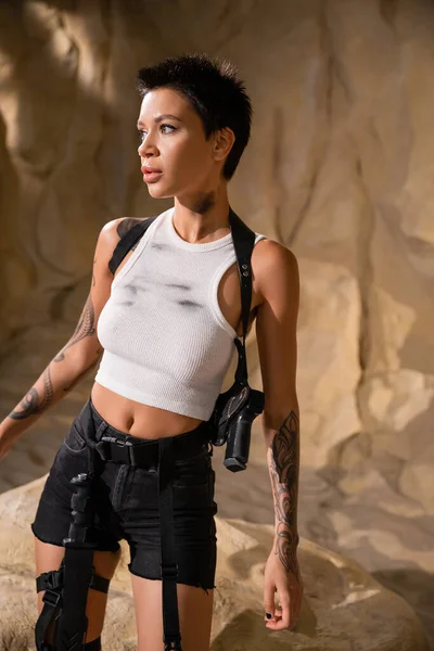Tattooed archaeologist in dirty outfit with gun in holster looking away in cave — Stock Photo