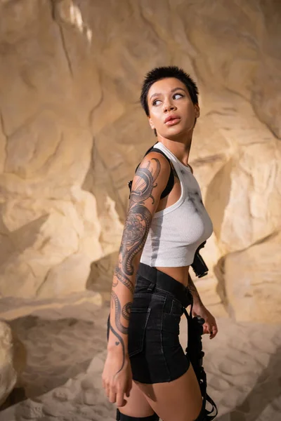Armed and tattooed archaeologist in sexy outfit looking away while walking in cave — Stock Photo