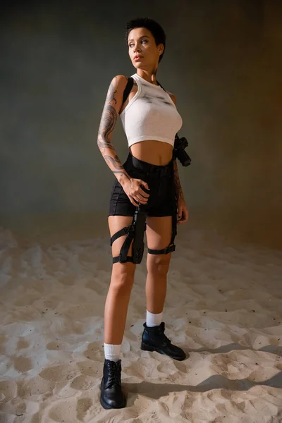 Full length of tattooed and armed archaeologist in dirty crop top looking away while standing on sand — Stock Photo