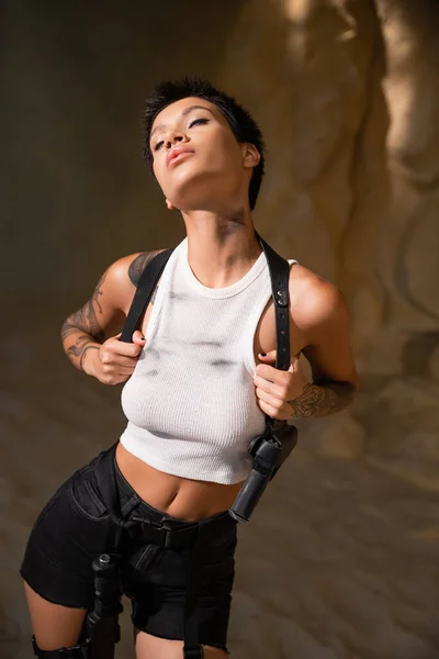 Sexy and armed archaeologist with closed eyes and dirty crop top adjusting holster with gun — Stock Photo
