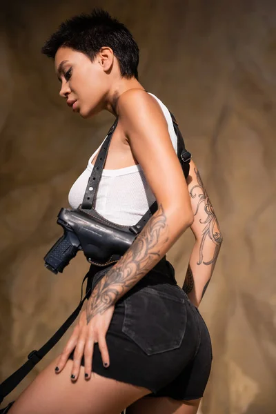 Tattooed archaeologist with short hair and crop top standing in holster with gun — Stock Photo