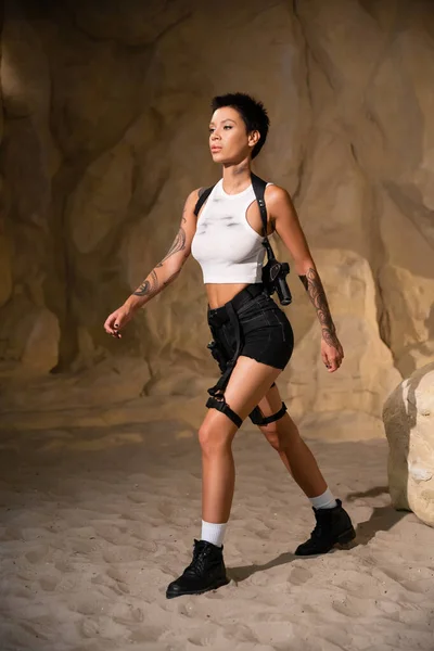 Full length of sexy and tattooed archaeologist with holster and gun walking on sand in cave — Stock Photo