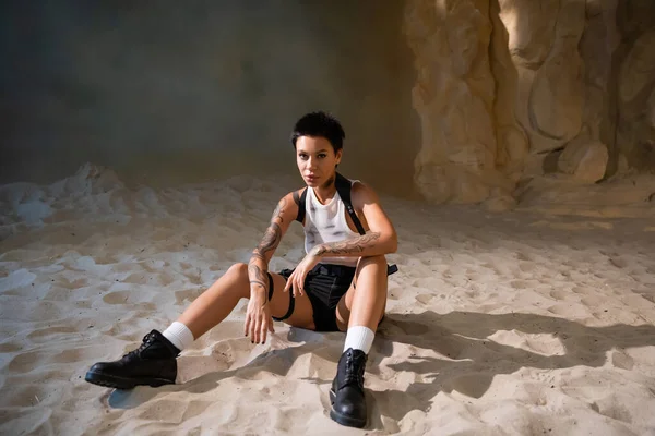 Longueur complète de tatoué archéologue dans sale crop top et shorts sexy assis sur le sable — Photo de stock