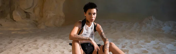 Archéologue tatoué et sexy en haut de culture sale et shorts assis sur le sable, bannière — Photo de stock