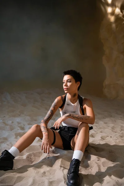 Full length of tattooed and sexy archaeologist in dirty crop top and sexy shorts sitting on sand in cave — Stock Photo