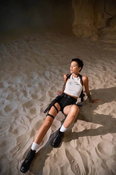 Vista ad alto angolo di giovane e sexy donna con tatuaggio e capelli corti seduta sulla sabbia — Foto stock