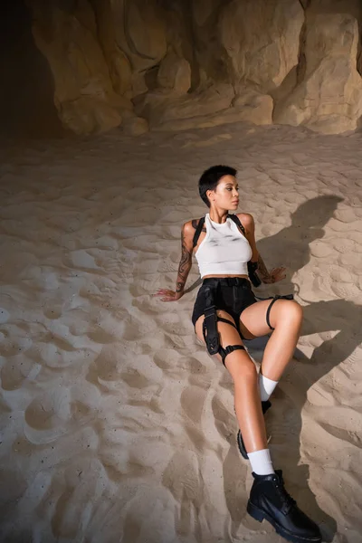 Высокий угол обзора вооруженной женщины с татуировкой и короткими волосами, сидящей на песке — стоковое фото