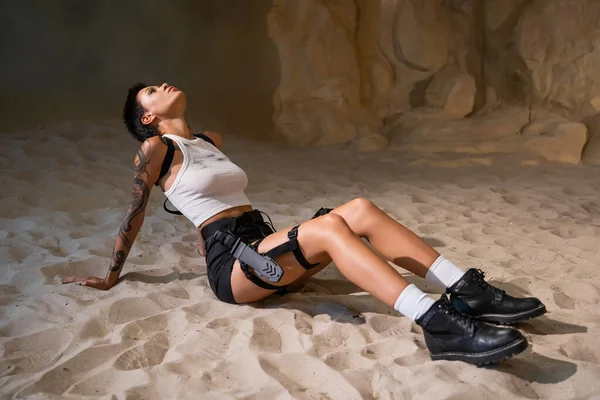 Tutta la lunghezza della giovane donna tatuata in abiti sexy sporchi seduta sulla sabbia — Foto stock