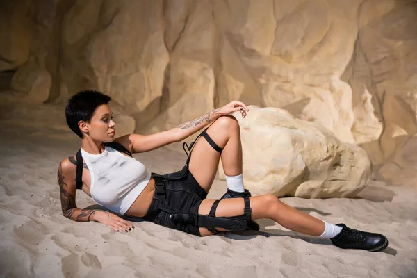 Longitud completa de arqueólogo tatuado en ropa sexy y funda tumbada en la arena en la cueva - foto de stock