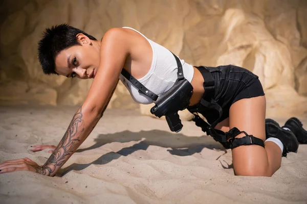Tatuato archeologo in abiti sexy e fondina strisciare sulla sabbia in grotta — Foto stock
