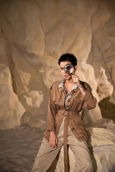 Jeune archéologue avec tatouage tenant loupe près du visage dans la grotte — Photo de stock